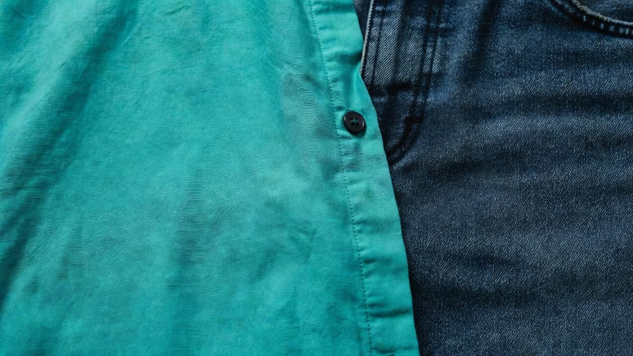 A close-up view displays the woven texture of a vibrant teal cotton shirt resting against the rugged, indigo-dyed weave of dark denim jeans under soft, clear daylight for high detail.